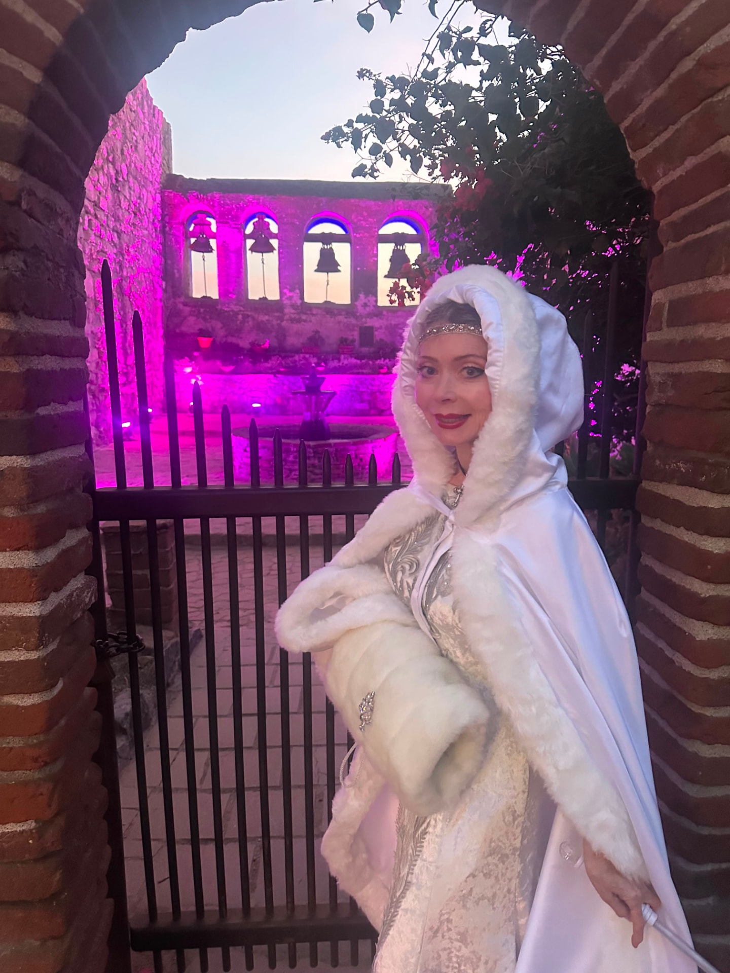 Dr. Marsha Long in a white fur-trimmed cloak beneath a brick archway at Mission San Juan Capistrano, the bell wall lit for Christmas