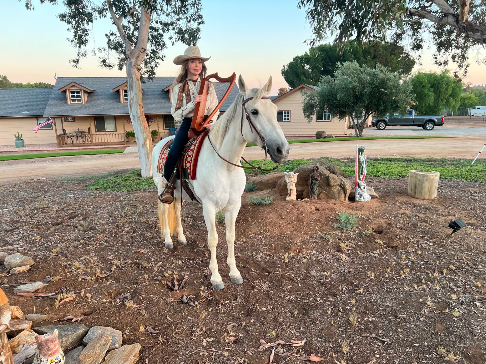Dr. Marsha Long in western dress and a cowboy hat, holding a lever harp while seated on her white horse Shasta at her San Diego ranch at dusk